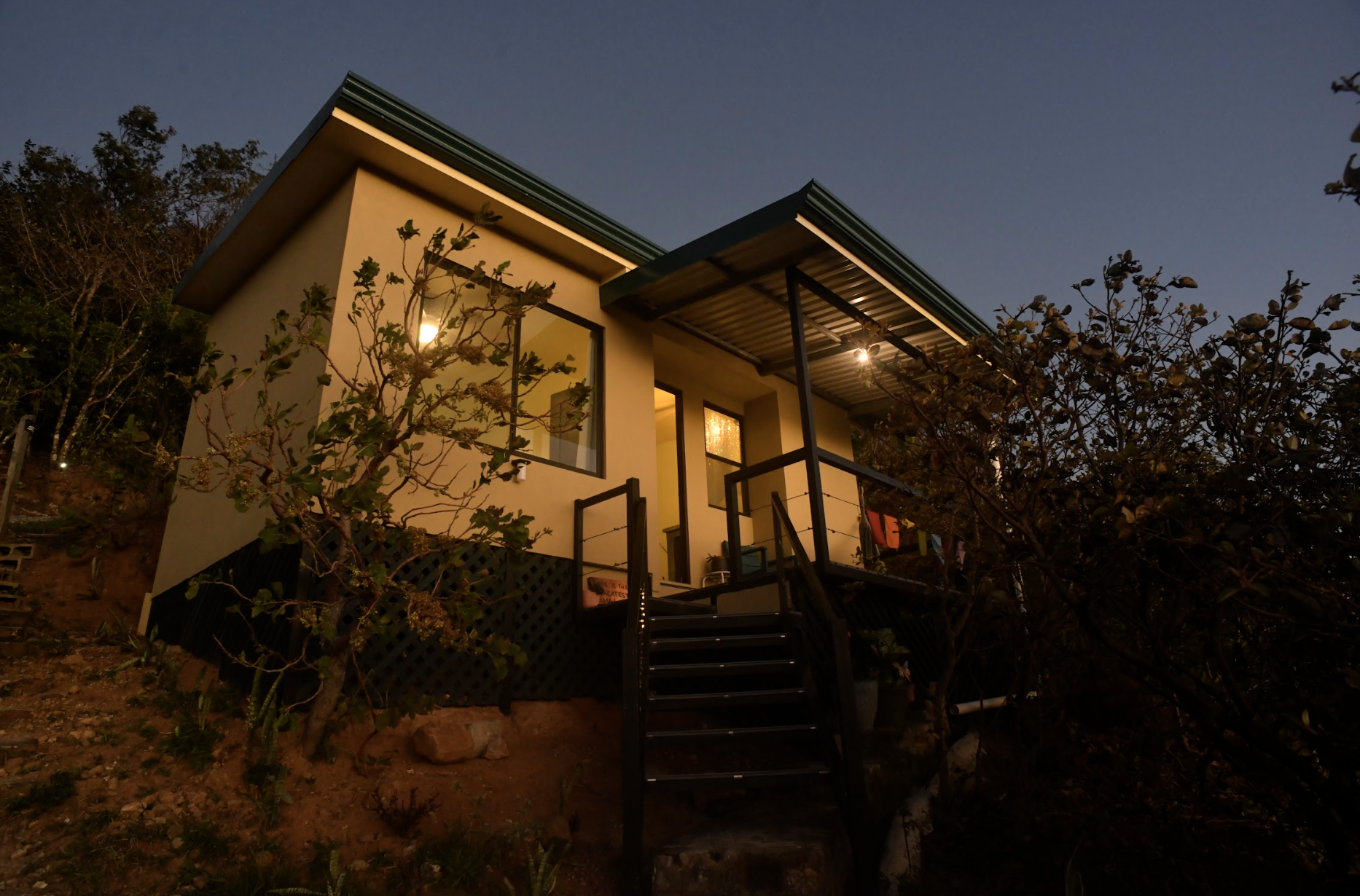 A modern house illuminated at dusk, situated on a hillside with trees and plants around, featuring a small outdoor staircase leading to an upper deck with a metal roof.
