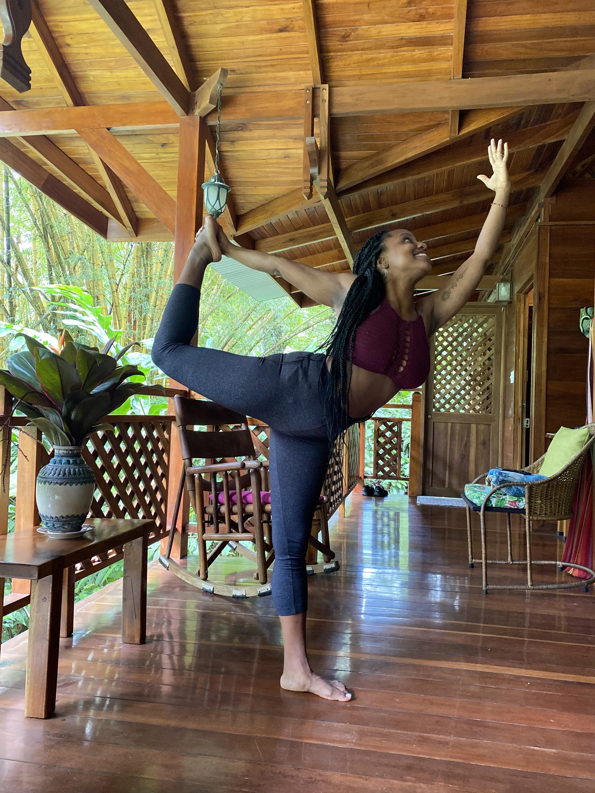 A woman practicing yoga on a wooden deck with a roof, surrounded by greenery, wearing a maroon top and dark leggings.