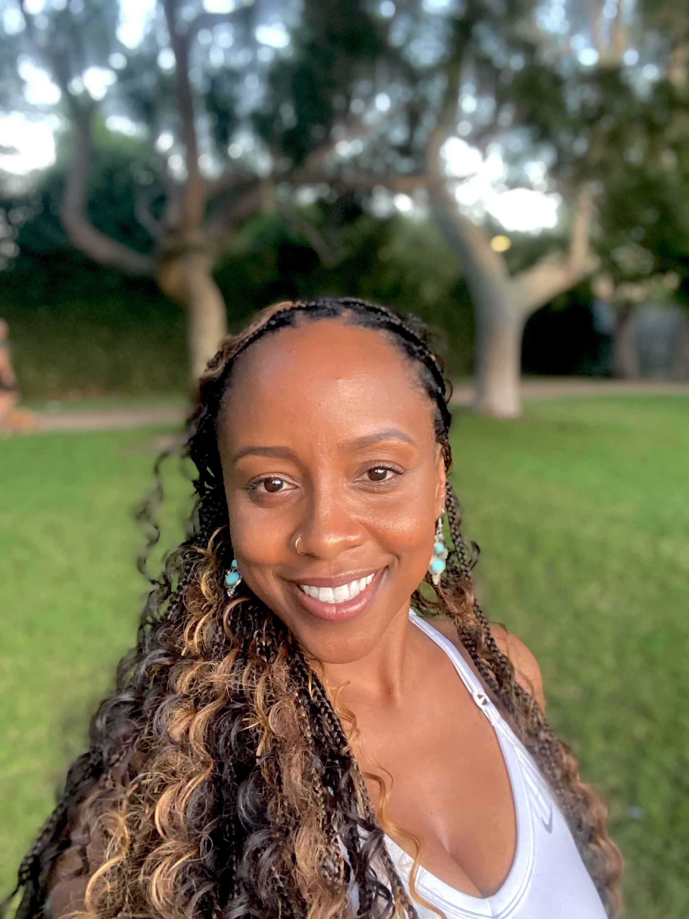 A smiling woman with long braided hair and earrings outdoors with trees and grass in the background.