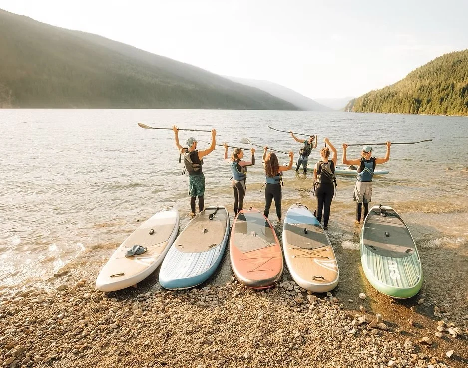 Five people holding paddles and preparing to start paddleboarding on a lake, with surfboards on the rocky shore and mountains in the background.