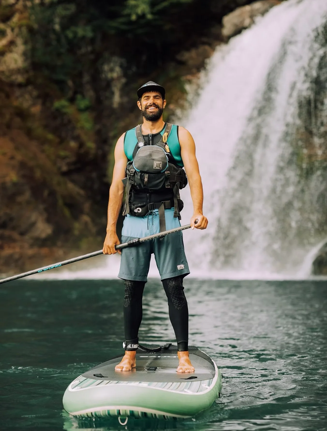 A man paddleboarding on a body of water with a waterfall in the background, wearing a cap, sleeveless shirt, shorts, and a life jacket.