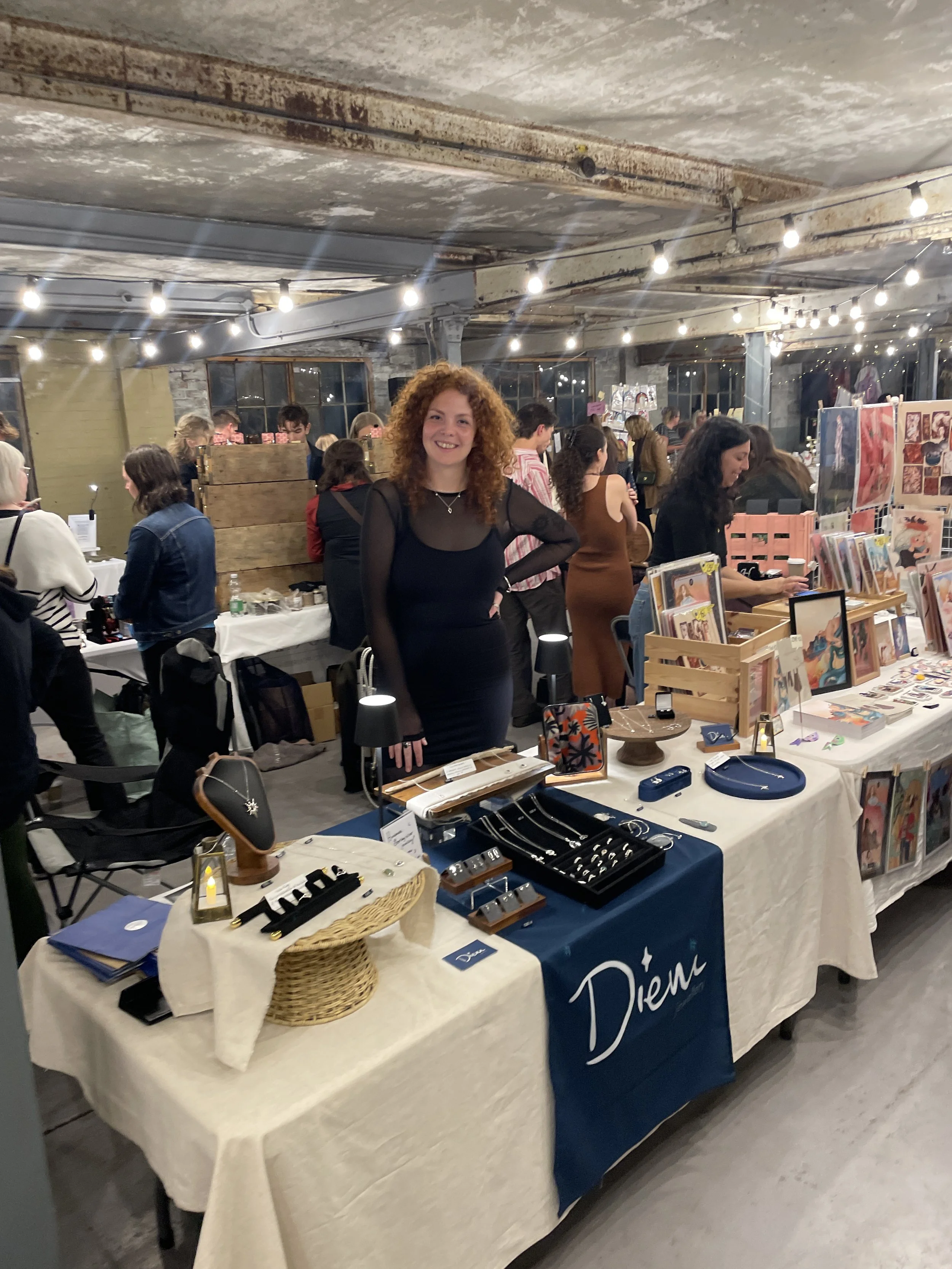 A woman with curly red hair smiling at a craft fair or market stall displaying jewelry and accessories. The stall has a blue tablecloth with white writing, and various jewelry pieces are showcased on display trays. Other people are browsing in the background inside an industrial-style space with string lights overhead.
