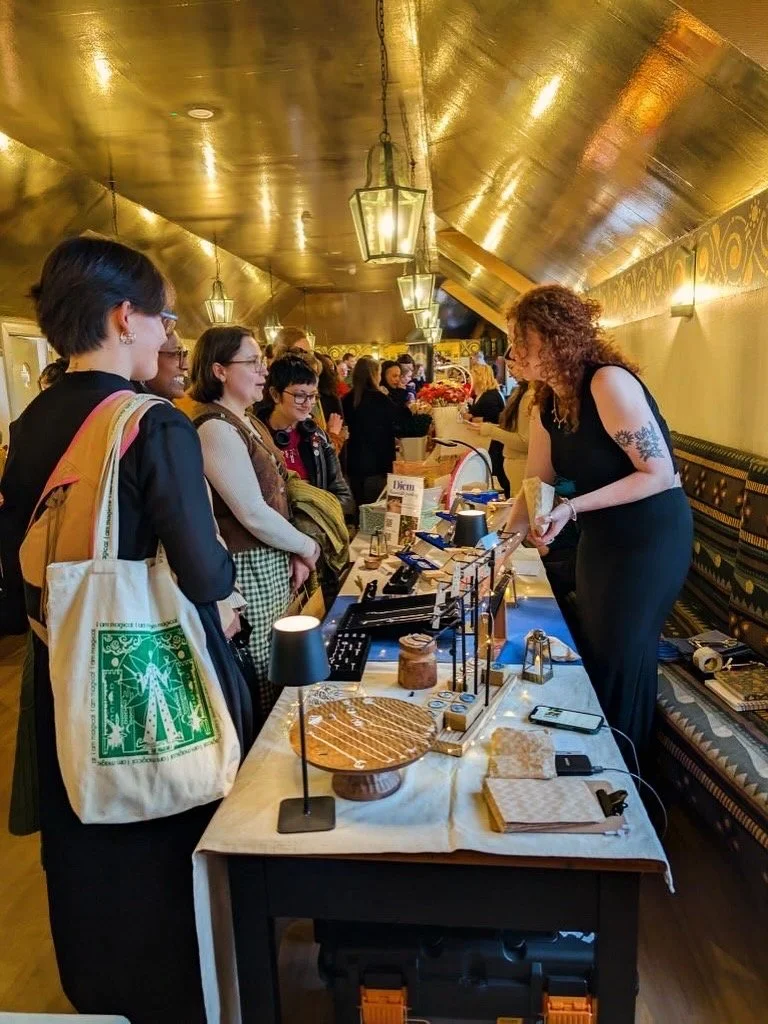 A group of women standing in line at a vendor booth in a warmly lit, gold-colored room. The vendor, a curly-haired woman in a black dress, is behind the table selling baked goods and other items. The table has baked desserts, small lamps, and display accessories. The room has a cozy, elegant atmosphere with a gold ceiling and patterned wall decor.