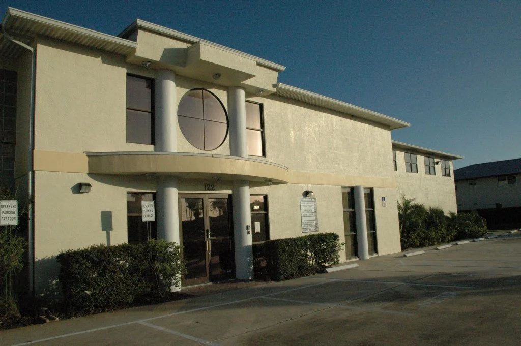 Exterior view of a beige commercial or office building with large cylindrical columns, a circular window, and a parking lot in front.
