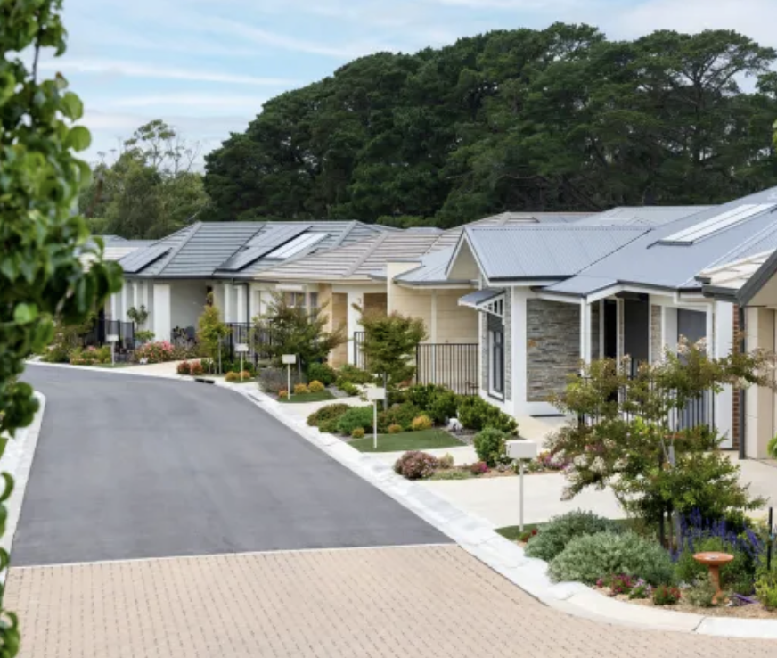 A row of modern single-family homes with landscaped front yards and a paved street, surrounded by trees.
