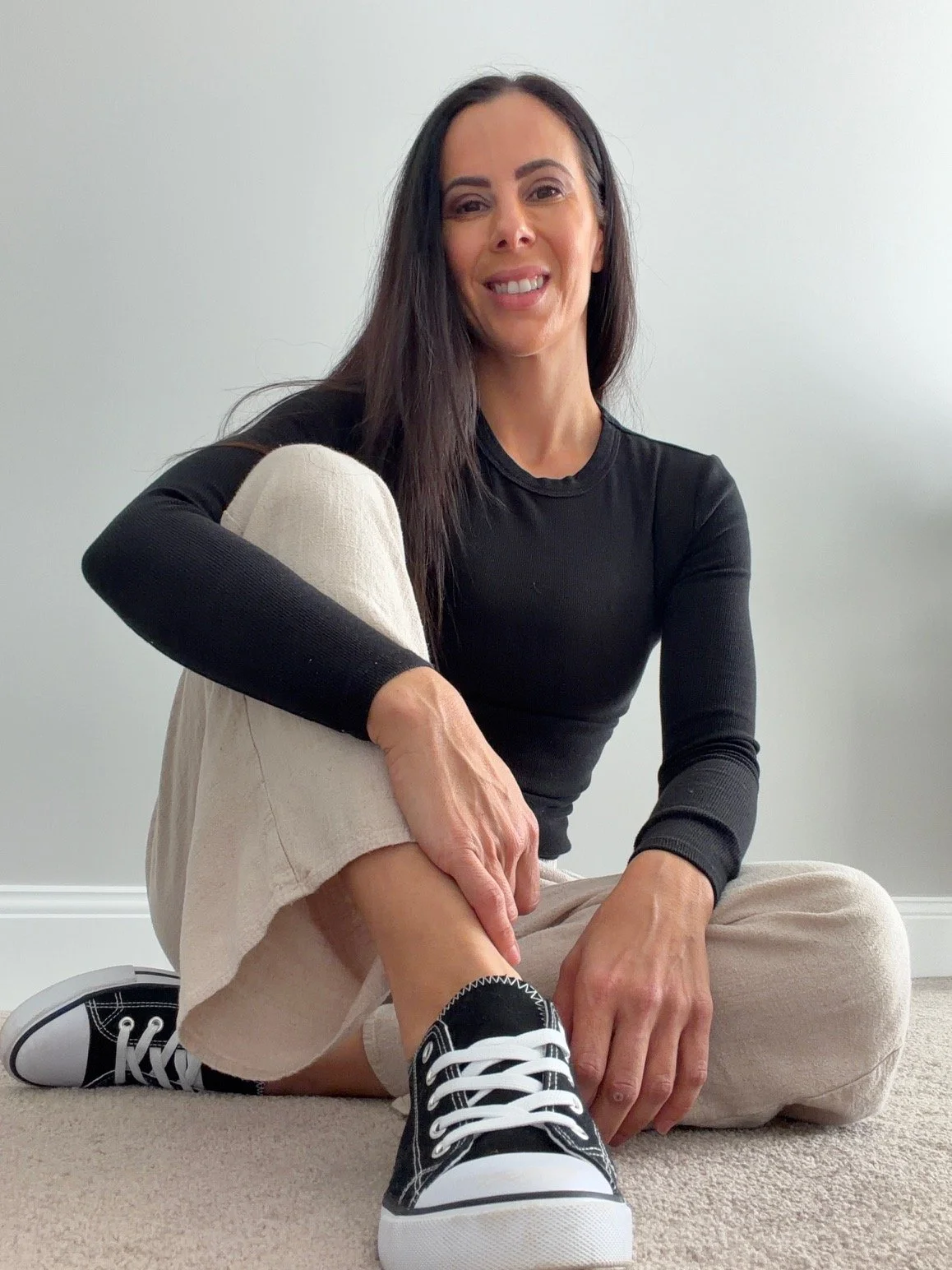 Woman with long dark hair wearing a black long sleeve shirt, beige pants, and black sneakers, sitting on the floor, holding her leg, smiling at the camera.
