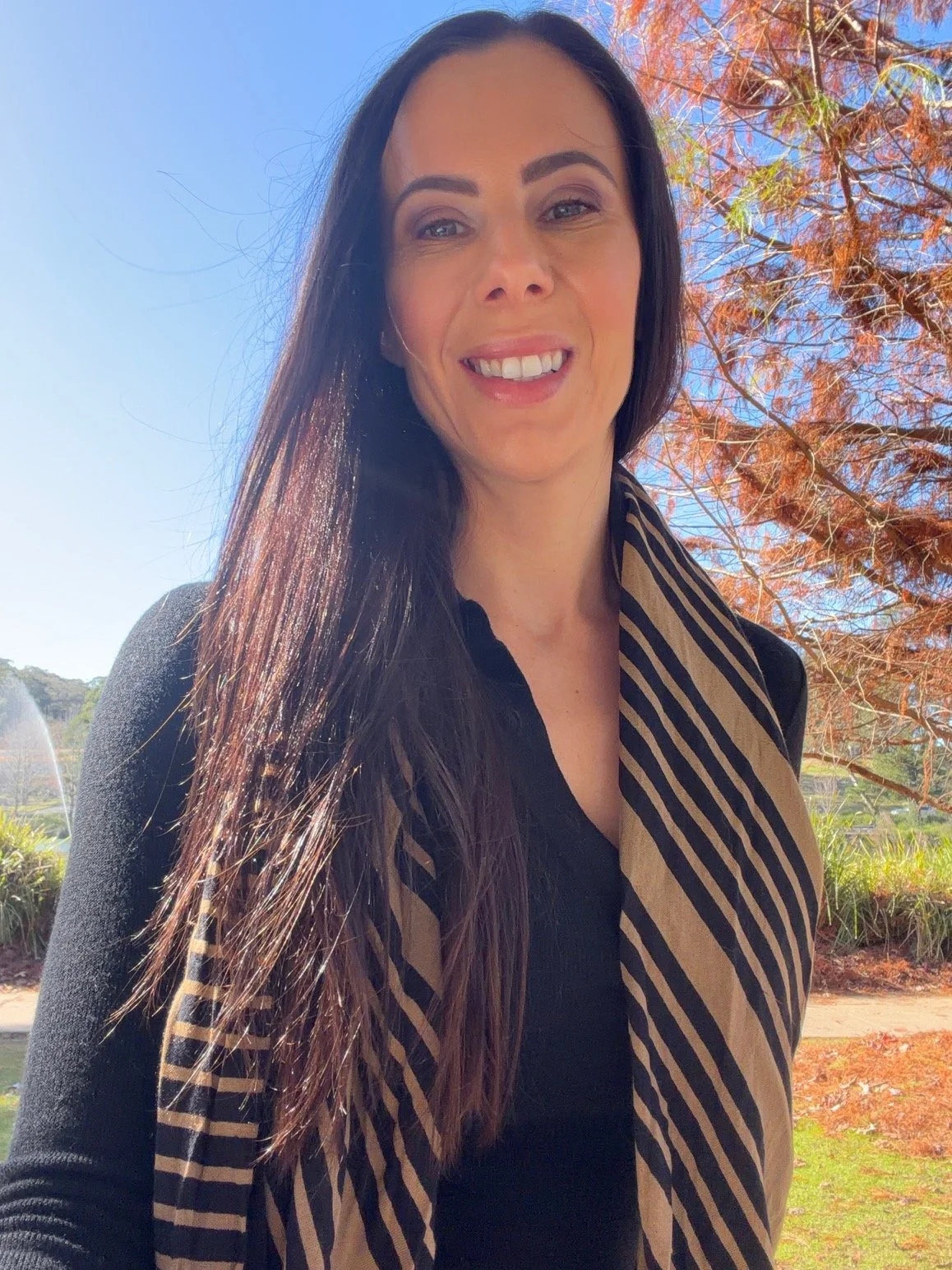 A woman with long dark hair smiling outdoors with autumn-colored trees and a fountain in the background.