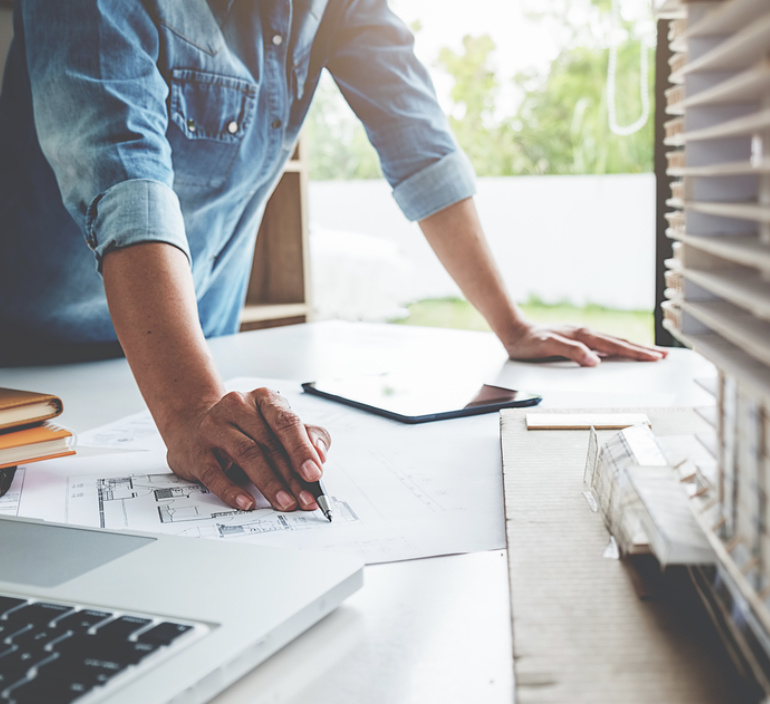 Person standing at a desk, working on architectural plans with a blueprint and a scale model, nearby a laptop and a tablet, in a bright room with large windows.