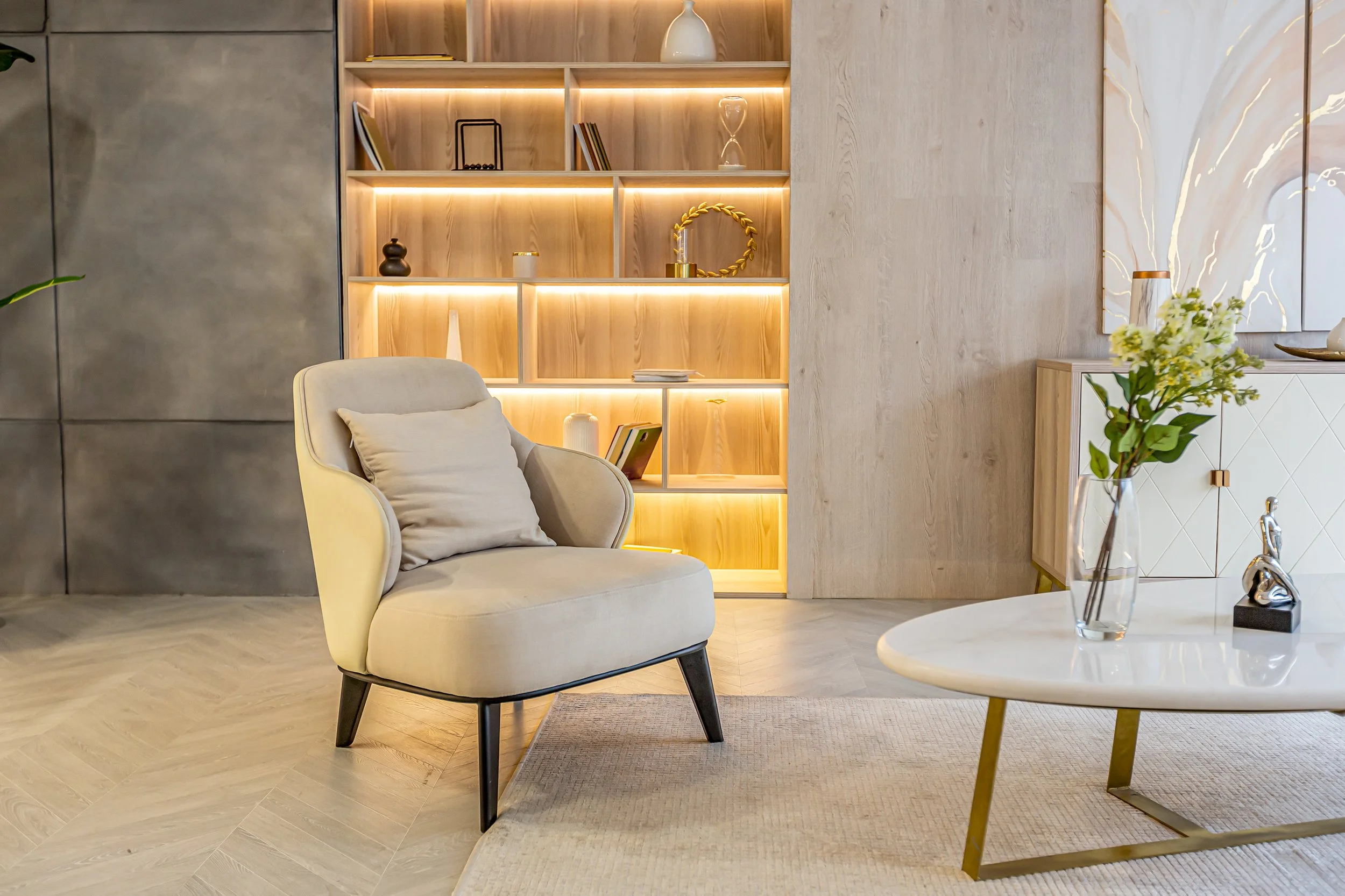 Modern living room with beige armchair, round white coffee table with a glass vase and flower arrangement, built-in wooden shelving with decorative items, and a sideboard with artwork.