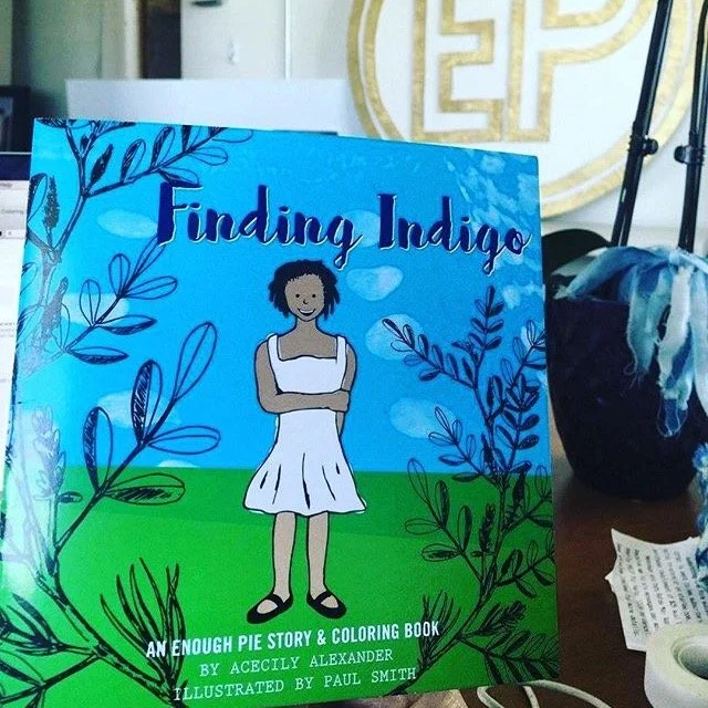 Cover of a children's storybook titled 'Finding Indigo' with an illustration of a girl in a white dress standing on green grass, surrounded by blue and black botanical drawings, with a featuring subtitle 'An Enough Pie Story & Coloring Book' and author mentions.