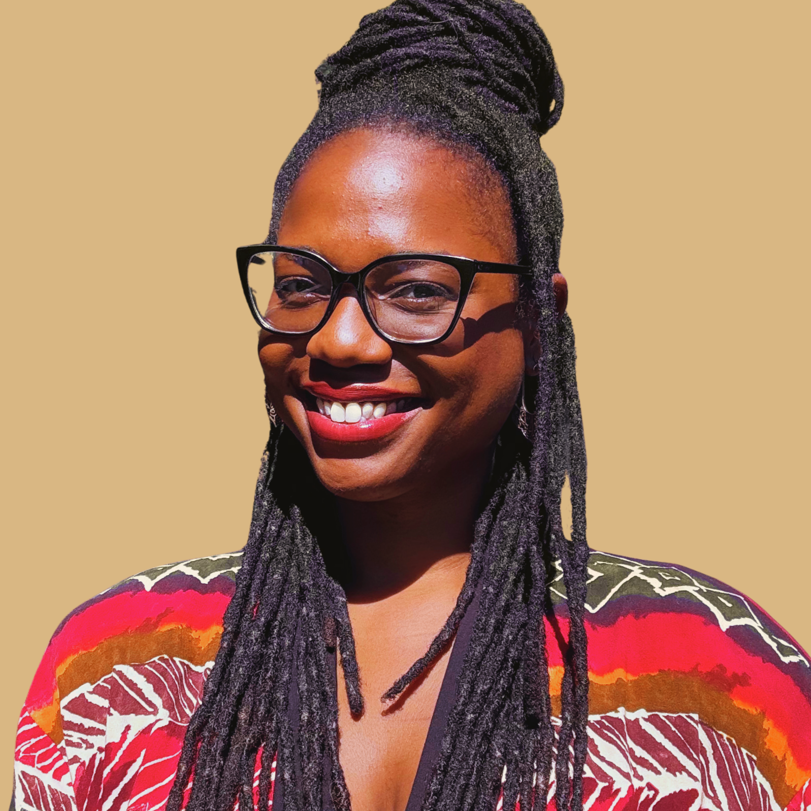A woman with dark skin, long braided hair, and glasses smiling, dressed in a vibrant red, orange, and purple patterned top, against a beige background.