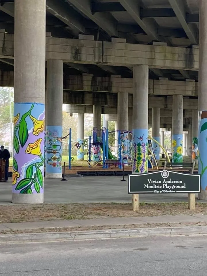 Colorful mural paintings on the concrete pillars of a playground under a bridge, with a sign reading Vivian Anderson Moultrie Playground, City of Charleston, in front of the playground.