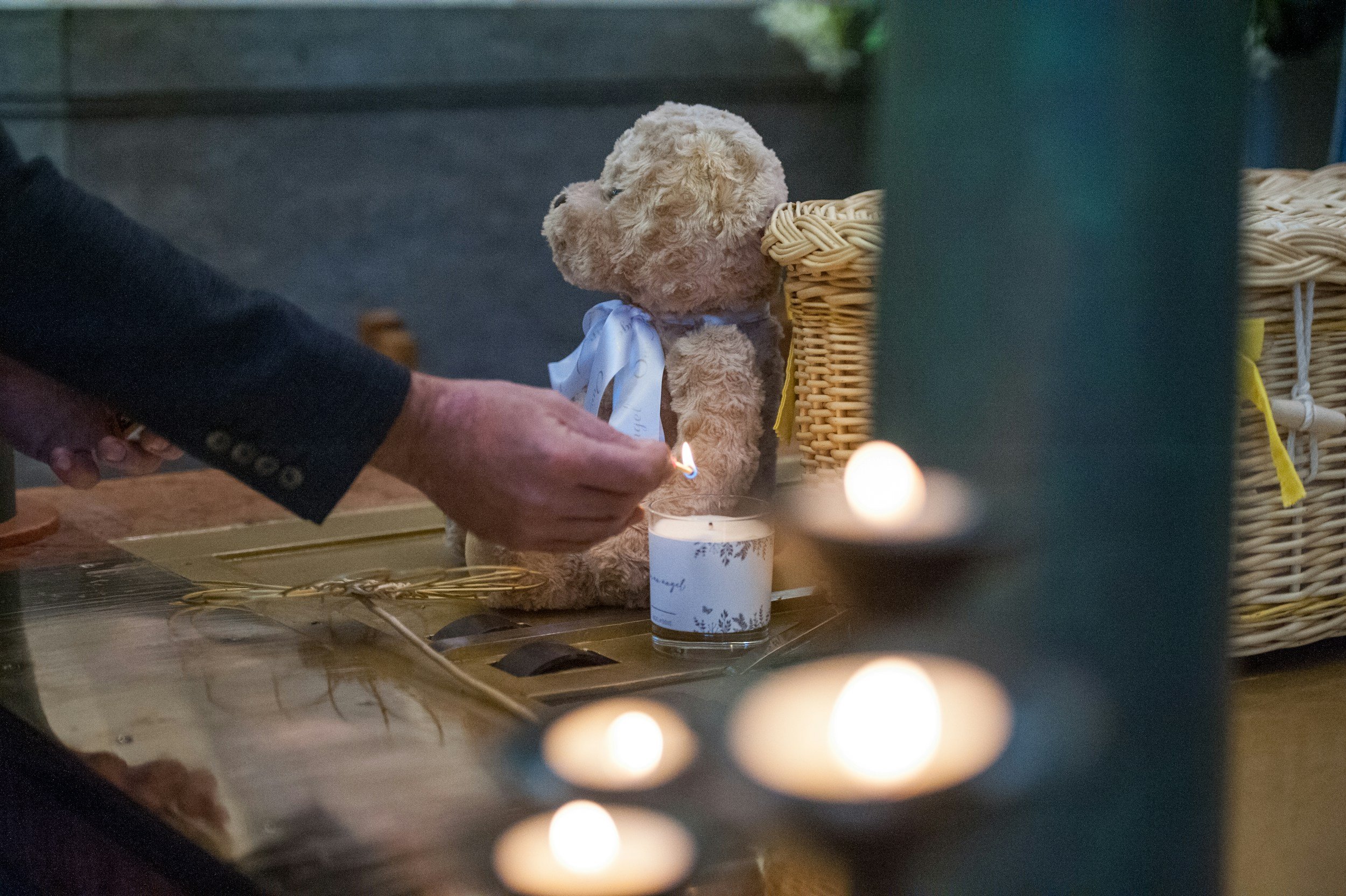 A teddy bear with a white ribbon sitting on a table, with candles and a person lighting a candle nearby.