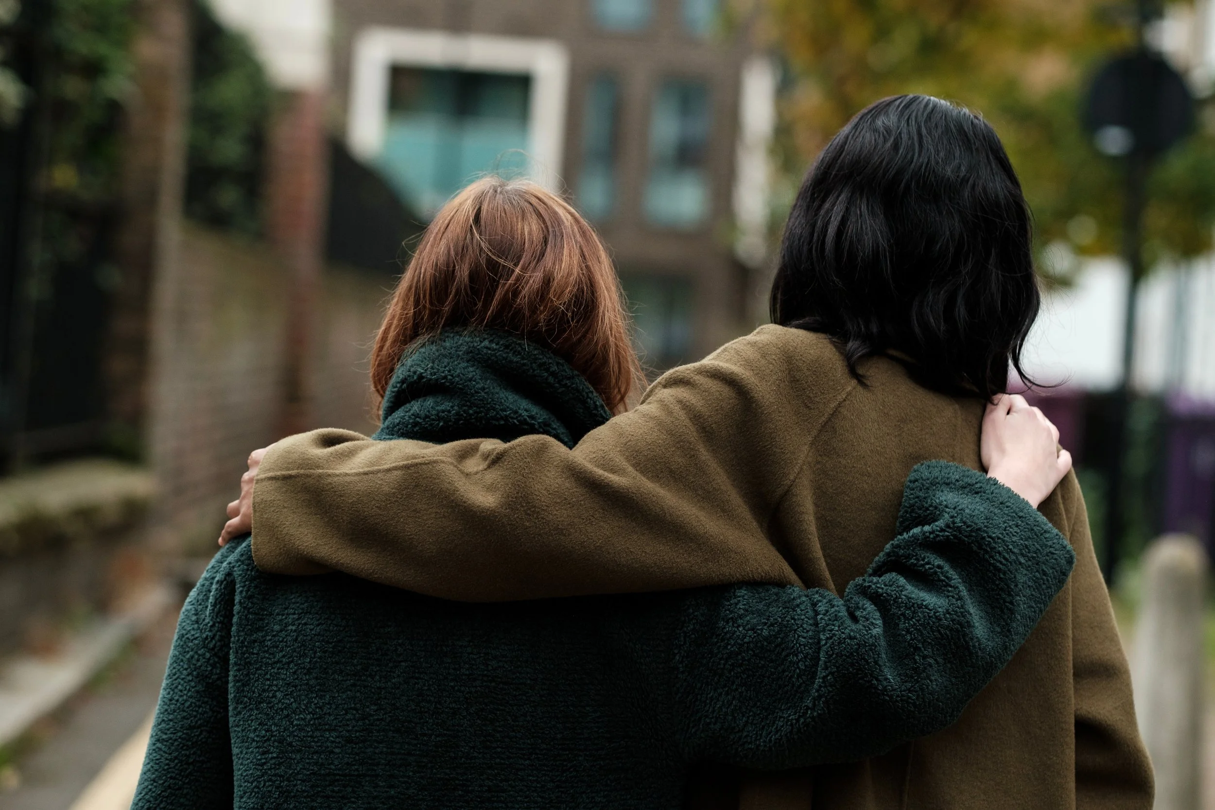 Two women hugging outdoors in a neighborhood setting with buildings and trees in the background.