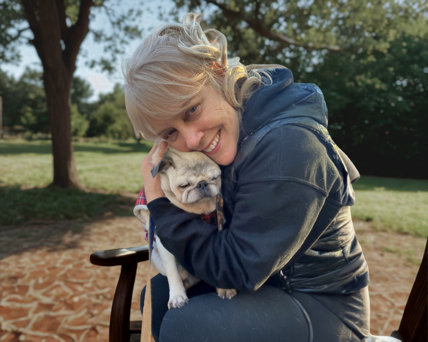 A woman hugging a small beige and white dog outdoors on a park bench, with trees in the background.