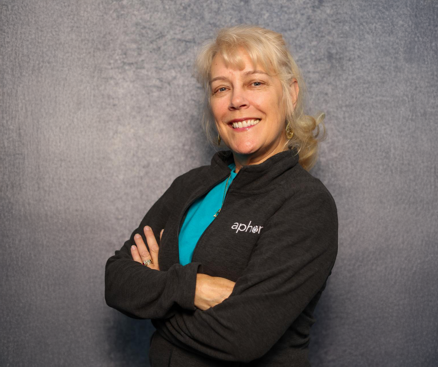 A woman with blonde hair smiling, wearing a black zip-up jacket with the logo 'aphor', standing with arms crossed against a gray textured background.