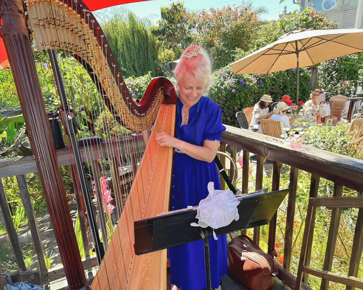 Woman with blonde hair playing a harp on a wooden deck in a garden with flowers and other people sitting under umbrellas.