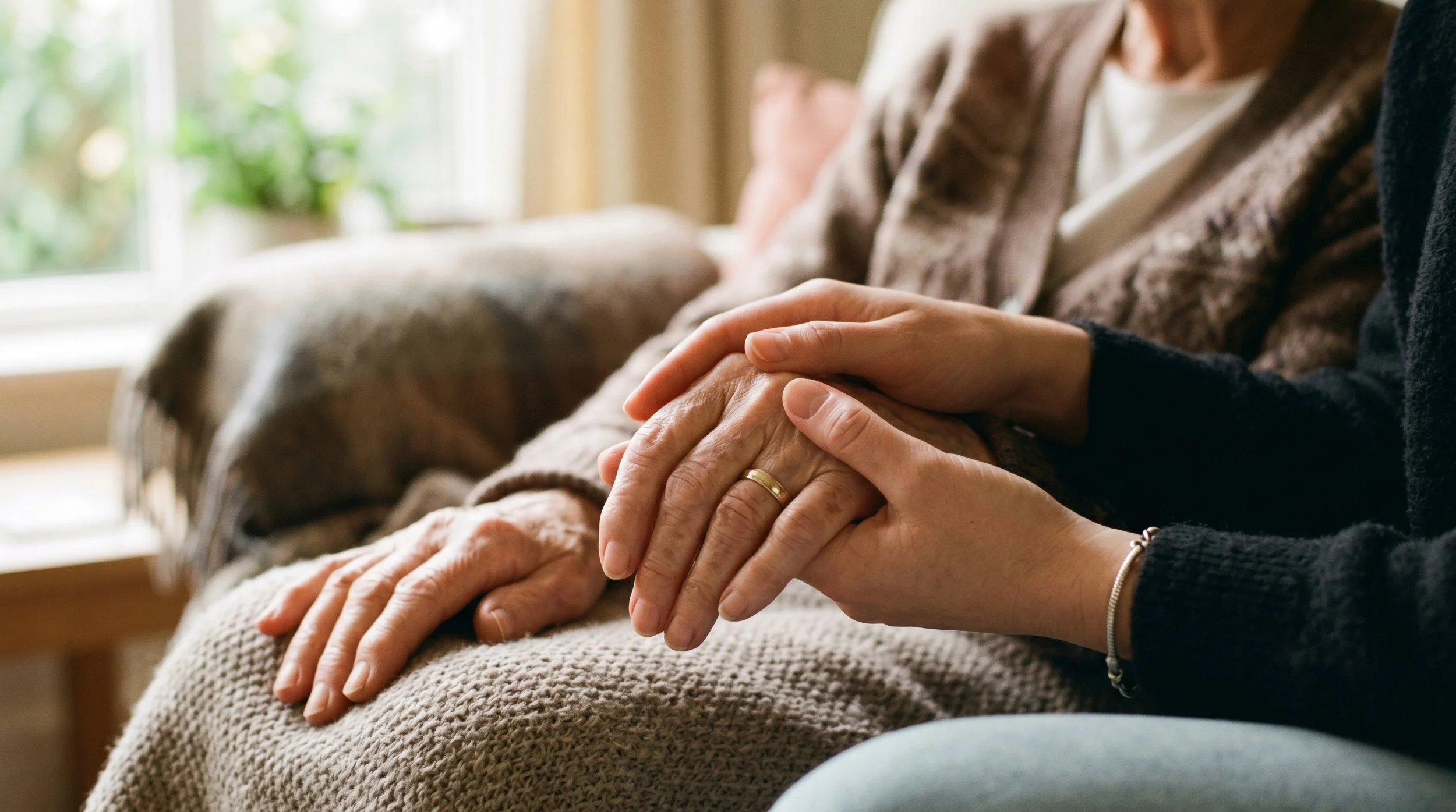 Two people, likely a caregiver and an elderly person, holding hands gently while sitting on a beige couch in a cozy, well-lit room with plants visible through a window.