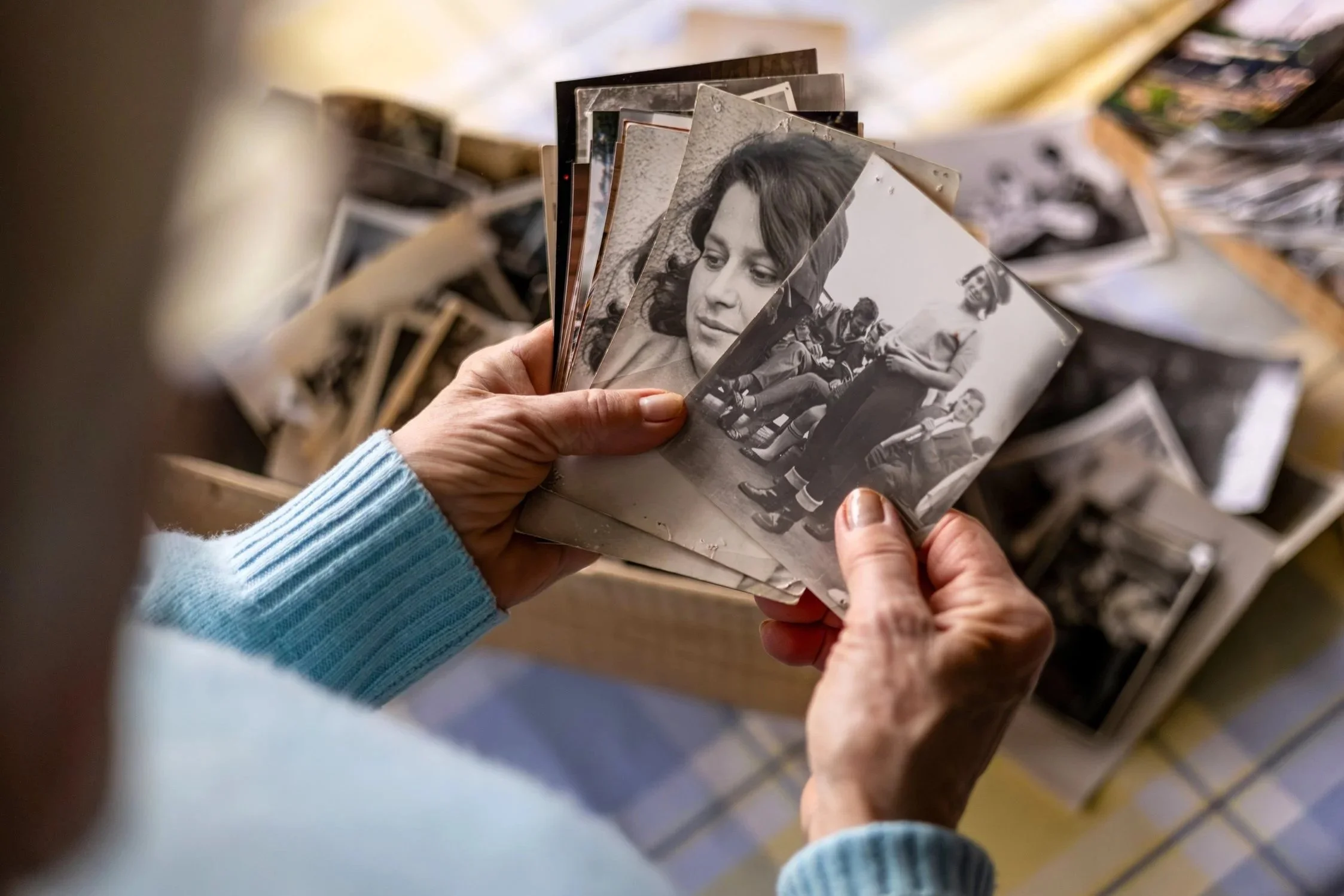 Person holding a stack of black-and-white photographs, with other photos scattered on a surface in the background.