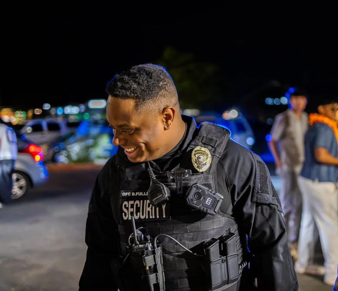 A smiling security officer wearing a black uniform and vest with equipment, standing outdoors at night with parked cars and blurred people in the background.