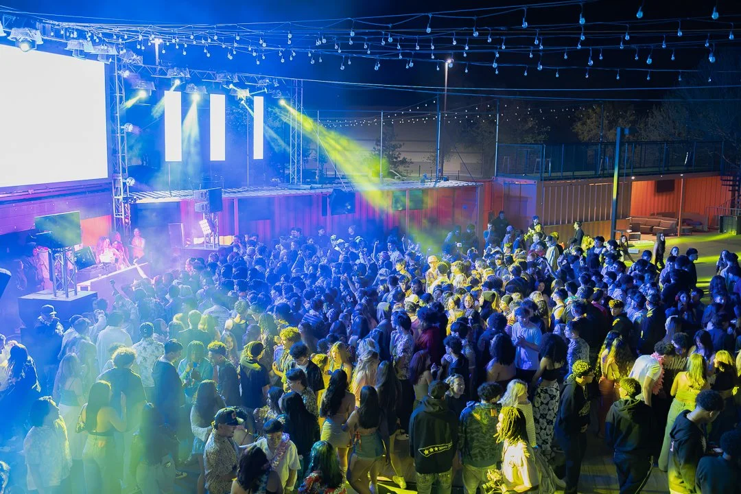 Crowd of people dancing and enjoying a live music event outdoors at night with stage, colorful lighting, and string lights overhead.