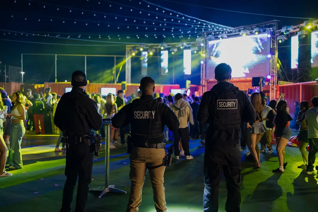Security personnel overseeing a crowd at a night outdoor concert or event with stage lights and screens in the background.
