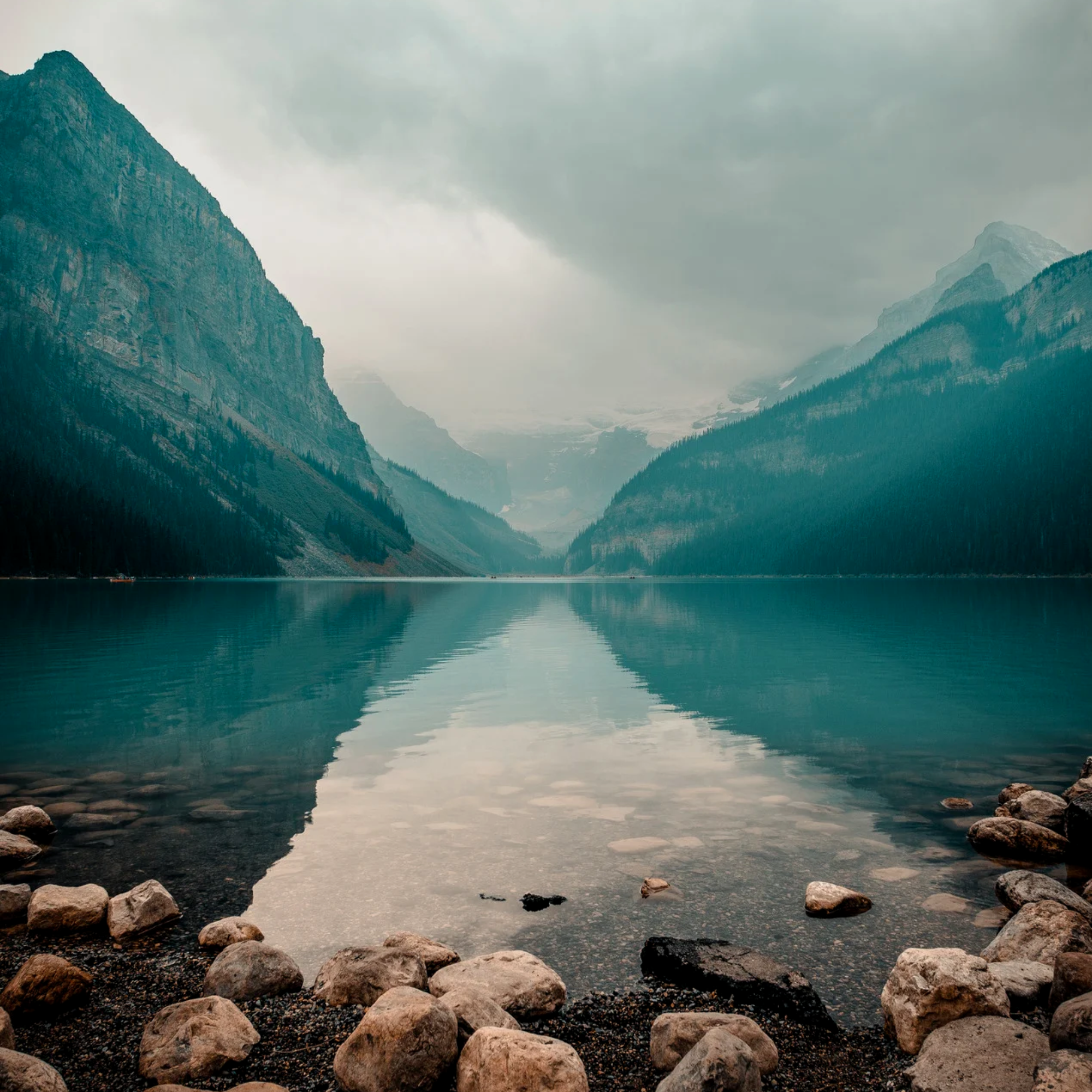 Calm mountain lake with rocky shore in the foreground, tall steep mountains on the left and right, snow-capped peaks in the distance, and cloudy sky reflected in the water.