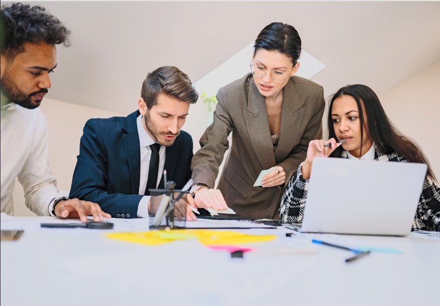 Five professionals gathered around a table in an office discussing documents and a laptop.