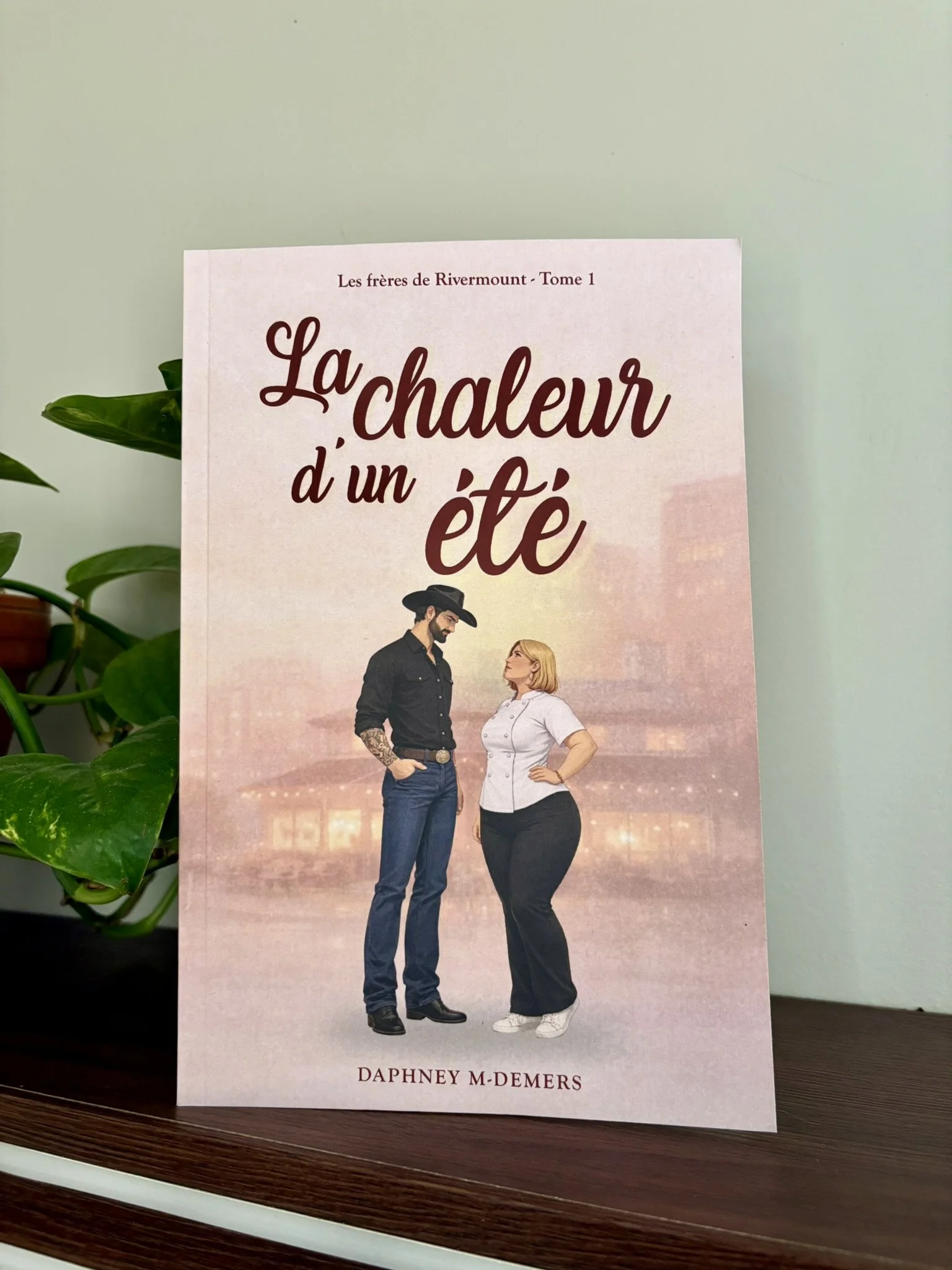 Couverture du livre 'La chaleur d’un été' de Daphney M. Demers, avec une illustration d’un homme portant un chapeau noir et une femme avec des cheveux blonds, face à face, contre un fond pastel avec des lumières de ville