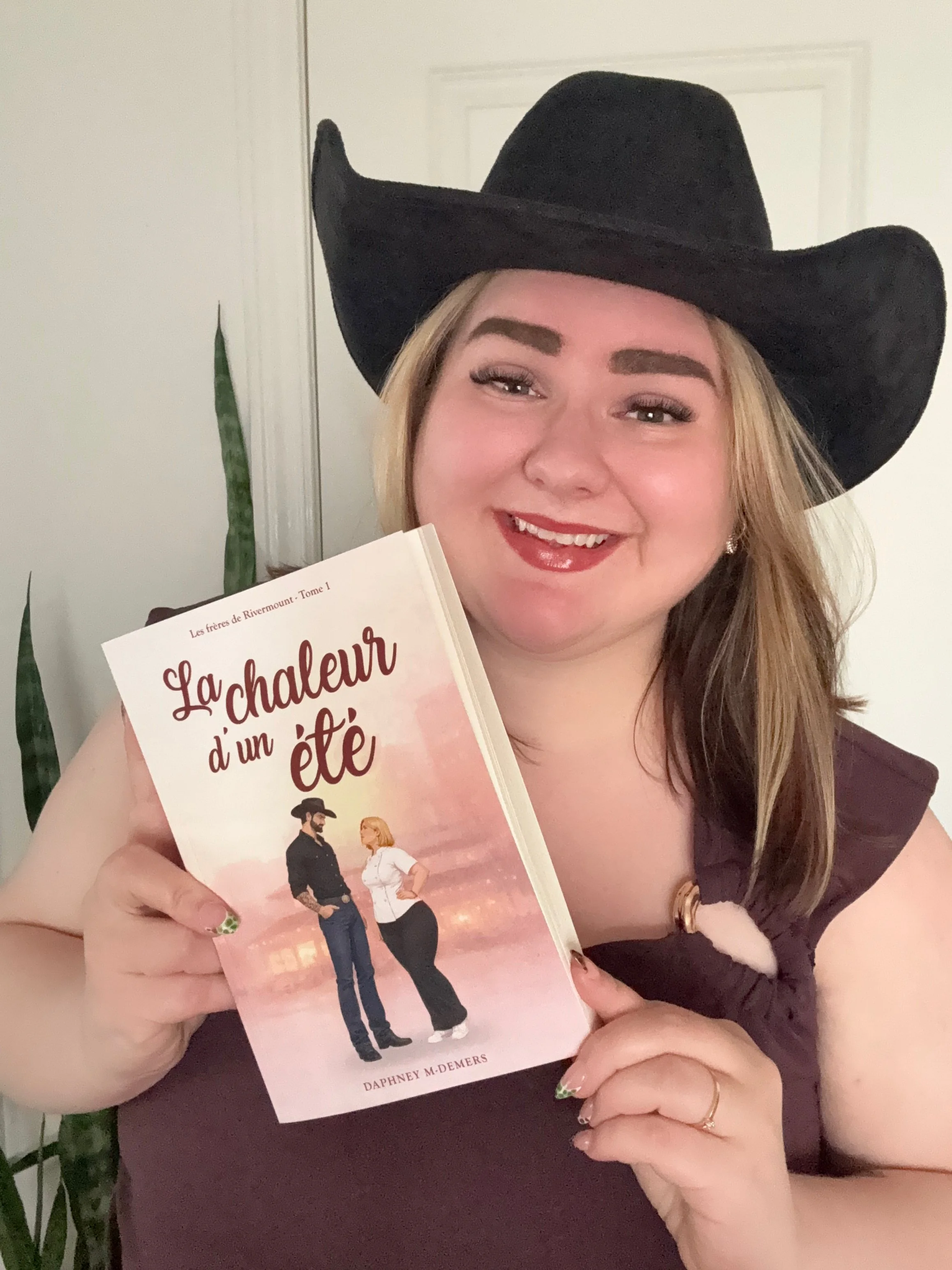 Une femme souriante porte un chapeau noir de cow-boy et tient un livre intitulé "La chaleur d'un été". Elle a des cheveux longs, maquillée avec du rouge à lèvres rouge et porte une robe maroon. Dans le fond, il y a des plantes vertes et un mur blanc.