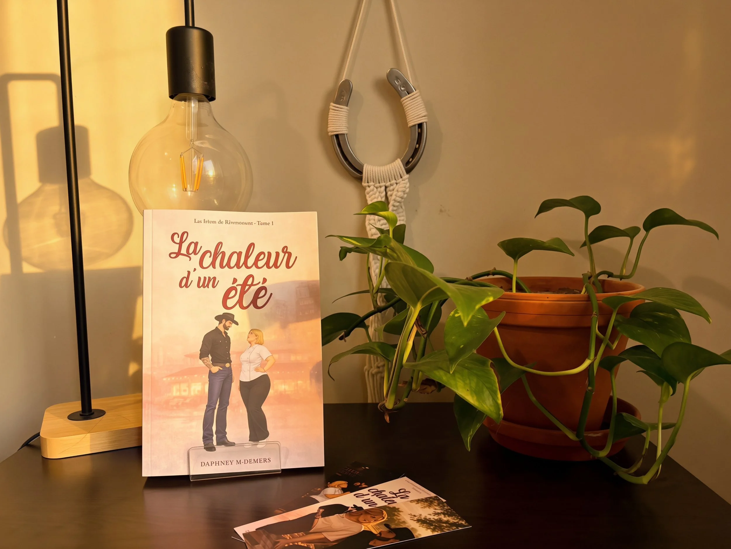 Une plante verte en pot, un livre avec une illustration de deux personnes parlant, une lampe à ampoule suspendue, un livre intitulé "La chaleur d’un été" posé sur une table, et un décor intérieur simple avec un mur beige.