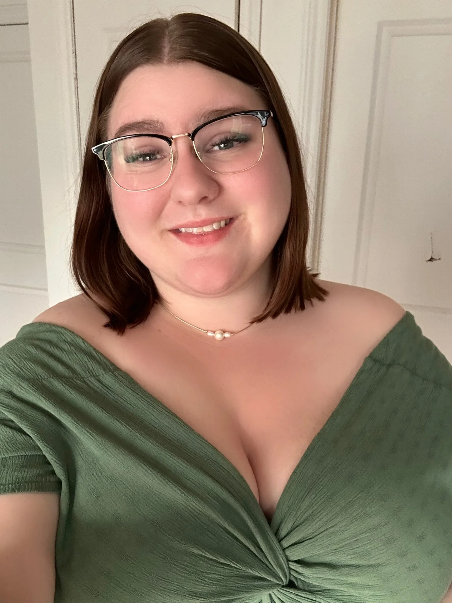 Jeune femme souriante portant des lunettes, une chaîne avec des perles et une robe verte, dans un intérieur avec des murs en bois peint en blanc.