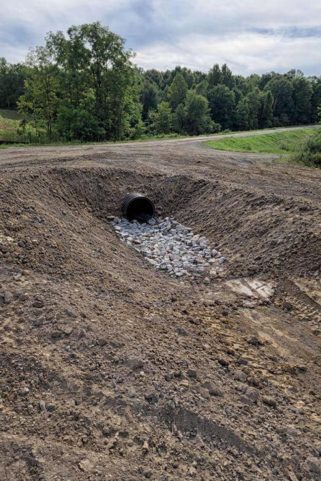 A drainage pipe protrudes from the ground, directing water and rocks through a channel in a dirt road construction site surrounded by trees and greenery.