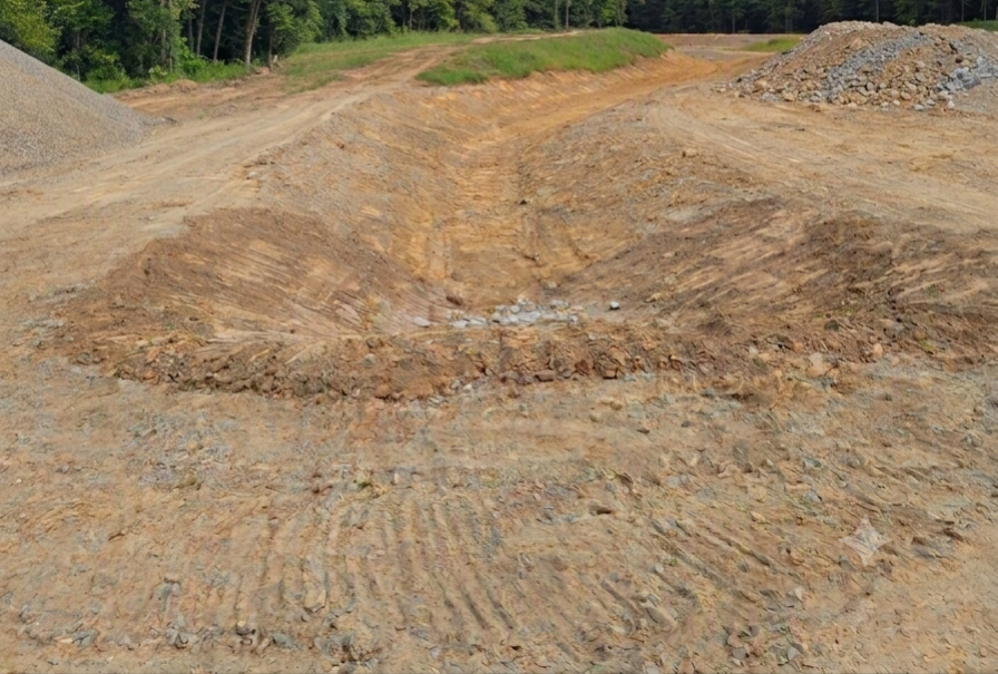 Excavated construction site with dirt mounds, tire tracks, and a forested background.