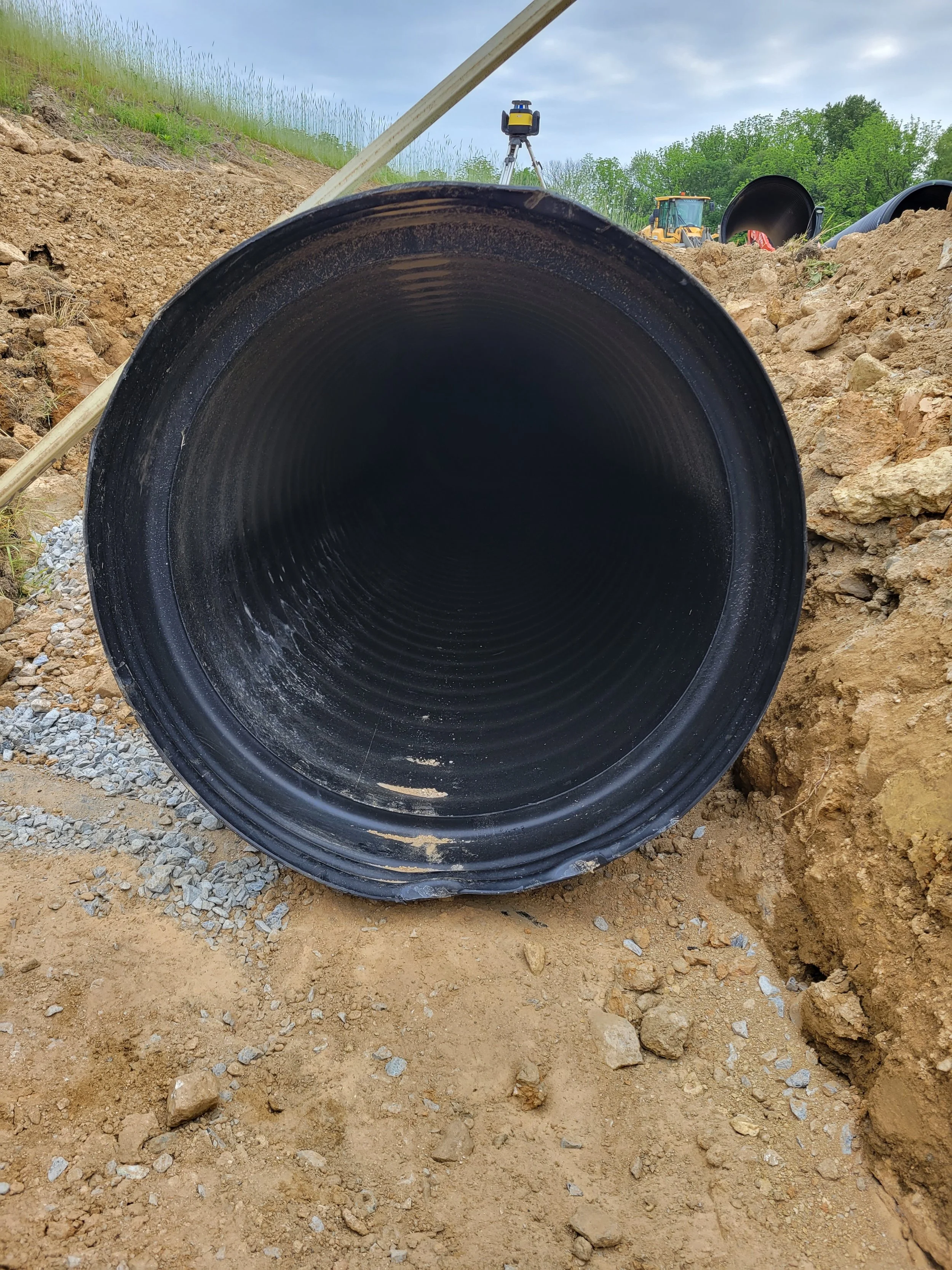 Close-up view of a large black pipe laying in a trench, with construction equipment and materials in the background.