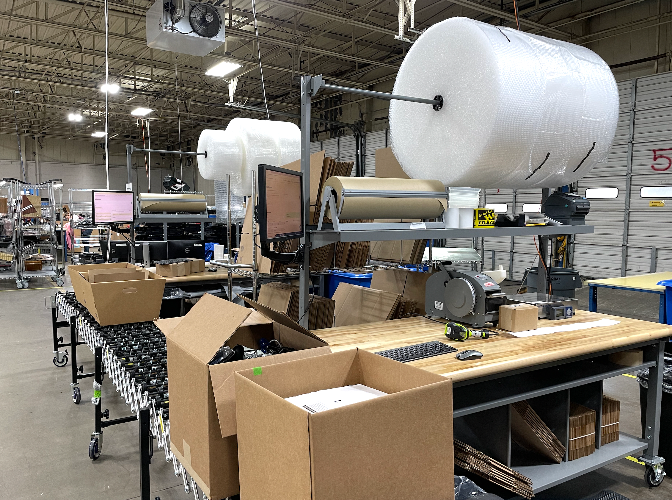 Interior of a warehouse or manufacturing facility with large rolls of bubble wrap on a conveyor system, surrounded by cardboard boxes, carts, and computers.