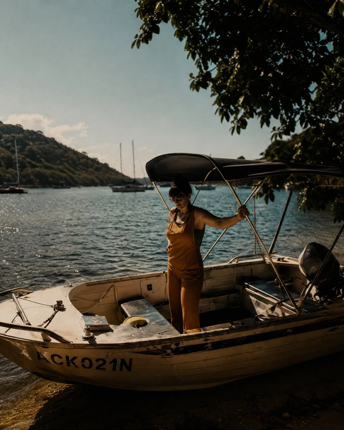 A woman standing in a small boat on the shore of a lake or bay, holding a canopy frame, with sailboats and a hillside in the background.