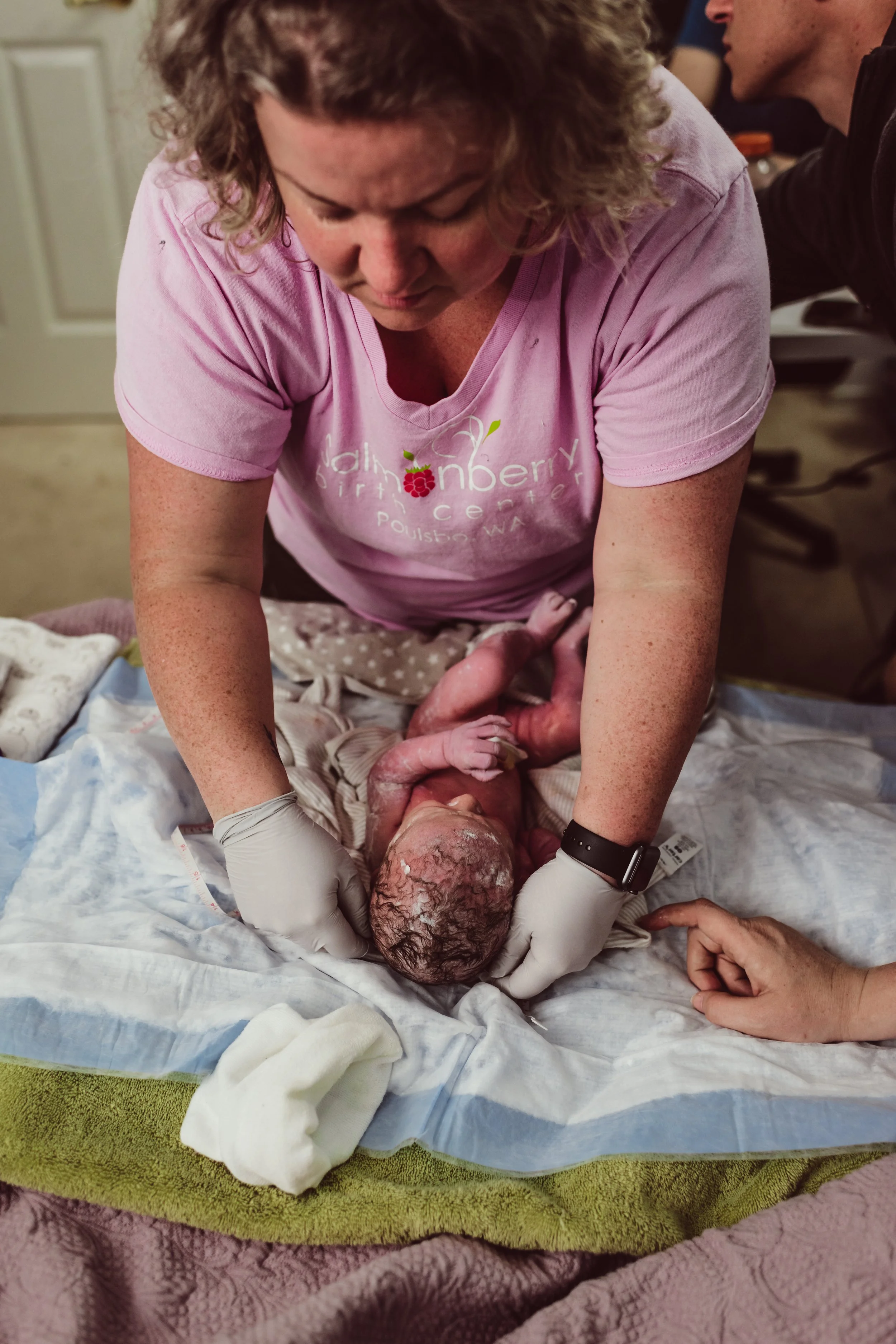A woman wearing a pink shirt and gloves is holding a newborn baby during childbirth in a hospital setting.