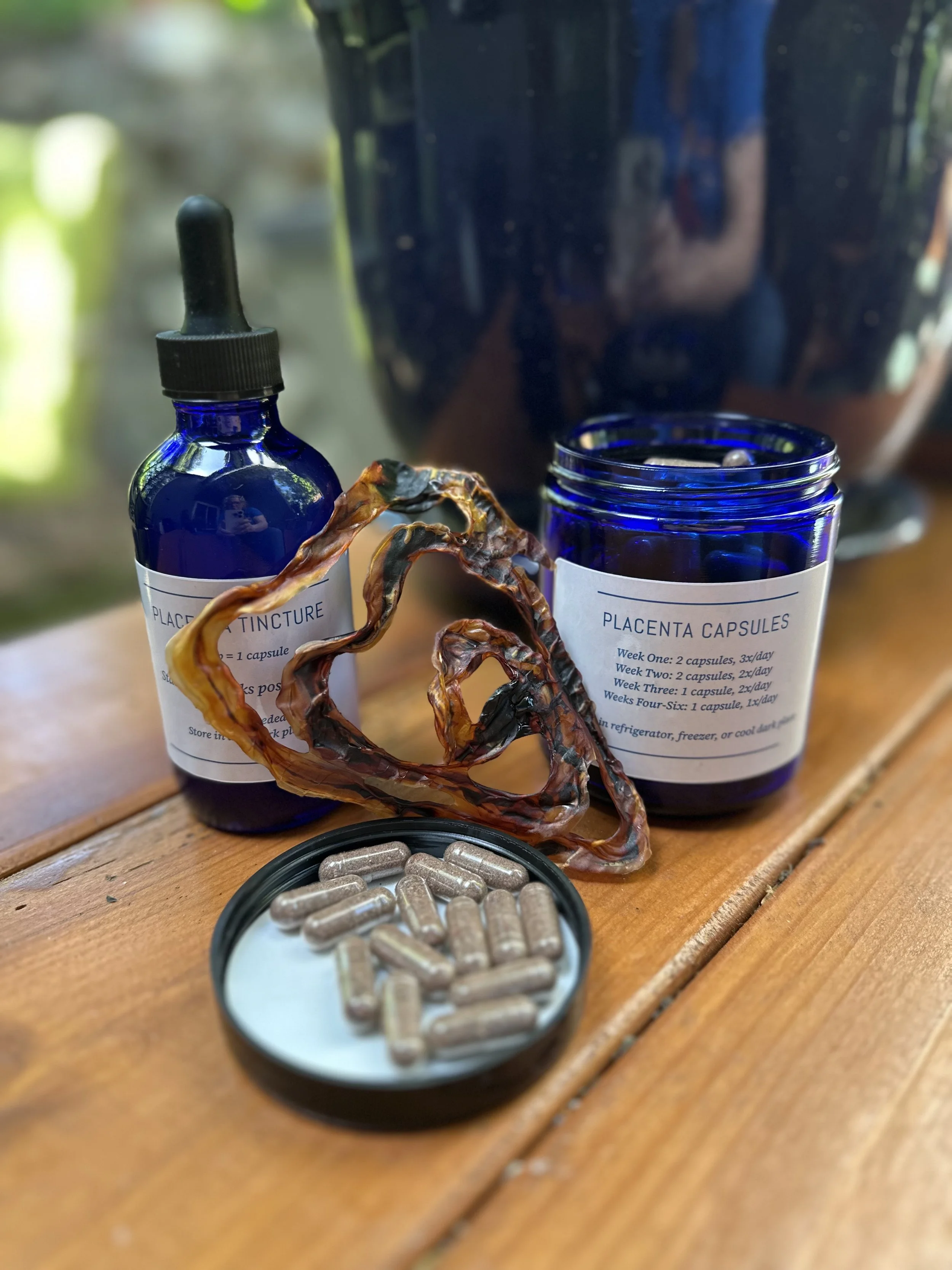 Blue bottles labeled 'Placenta Capsules' and 'Placenta Tincture'. Placenta capsules on a small dish on a wooden surface and example of a cord keepsake.