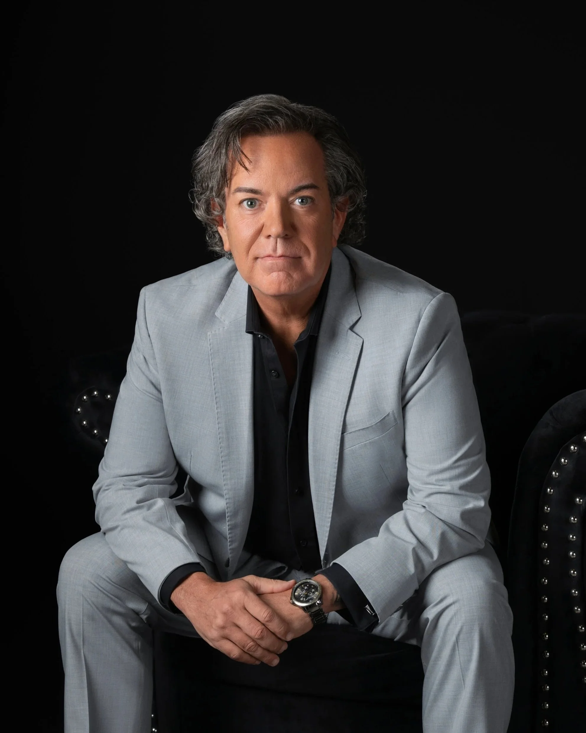 Jerod Arreguini, founder of The Art of Presenting, seated in a light gray suit and black shirt against a black background.