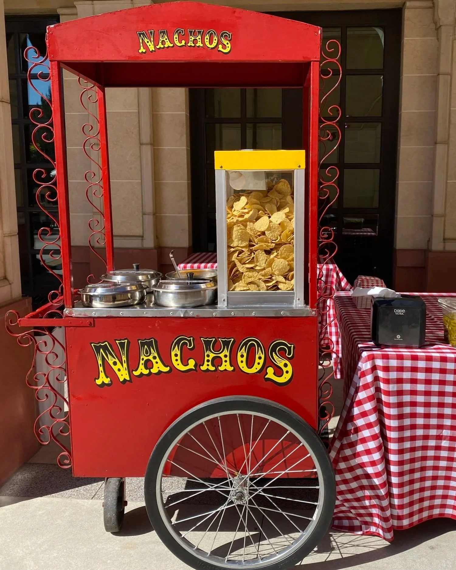 A red nacho and snack cart with decorative scrollwork, featuring a large sign that says 'Nachos'. The cart has a clear container filled with potato chips and metal containers likely holding toppings or condiments. There is a tray covered with a checkered red and white tablecloth next to the cart.