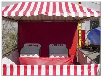 A carnival game stall with red and white striped canopy and backdrop, featuring two white beds with red and black lettering, set outdoors.