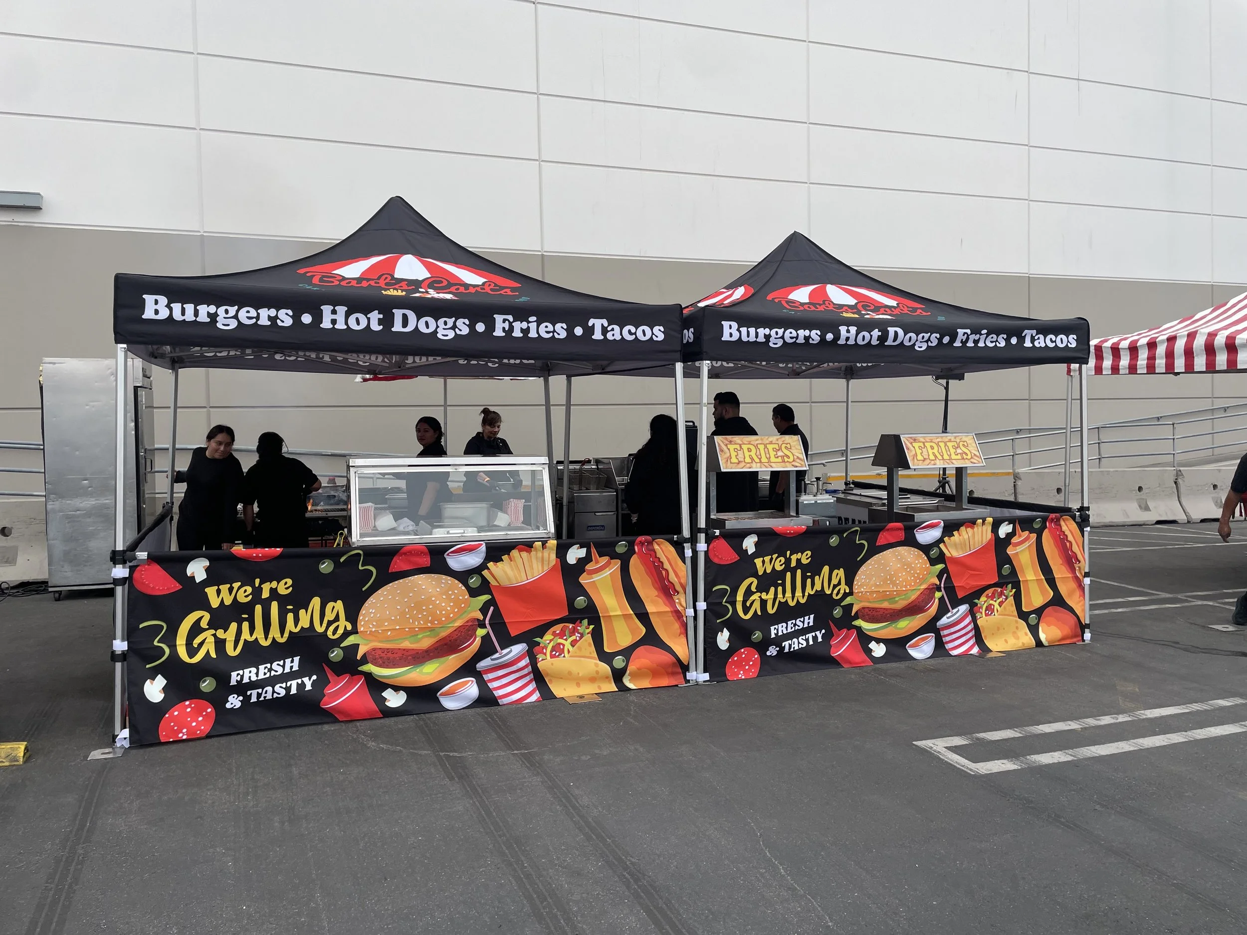 Outdoor food stand with black canopy advertising burgers, hot dogs, fries, and tacos. The front of the stand features colorful graphics of burgers, fries, and condiments, with the text "We're Grillin' Fresh & Tasty." There are several people working 
