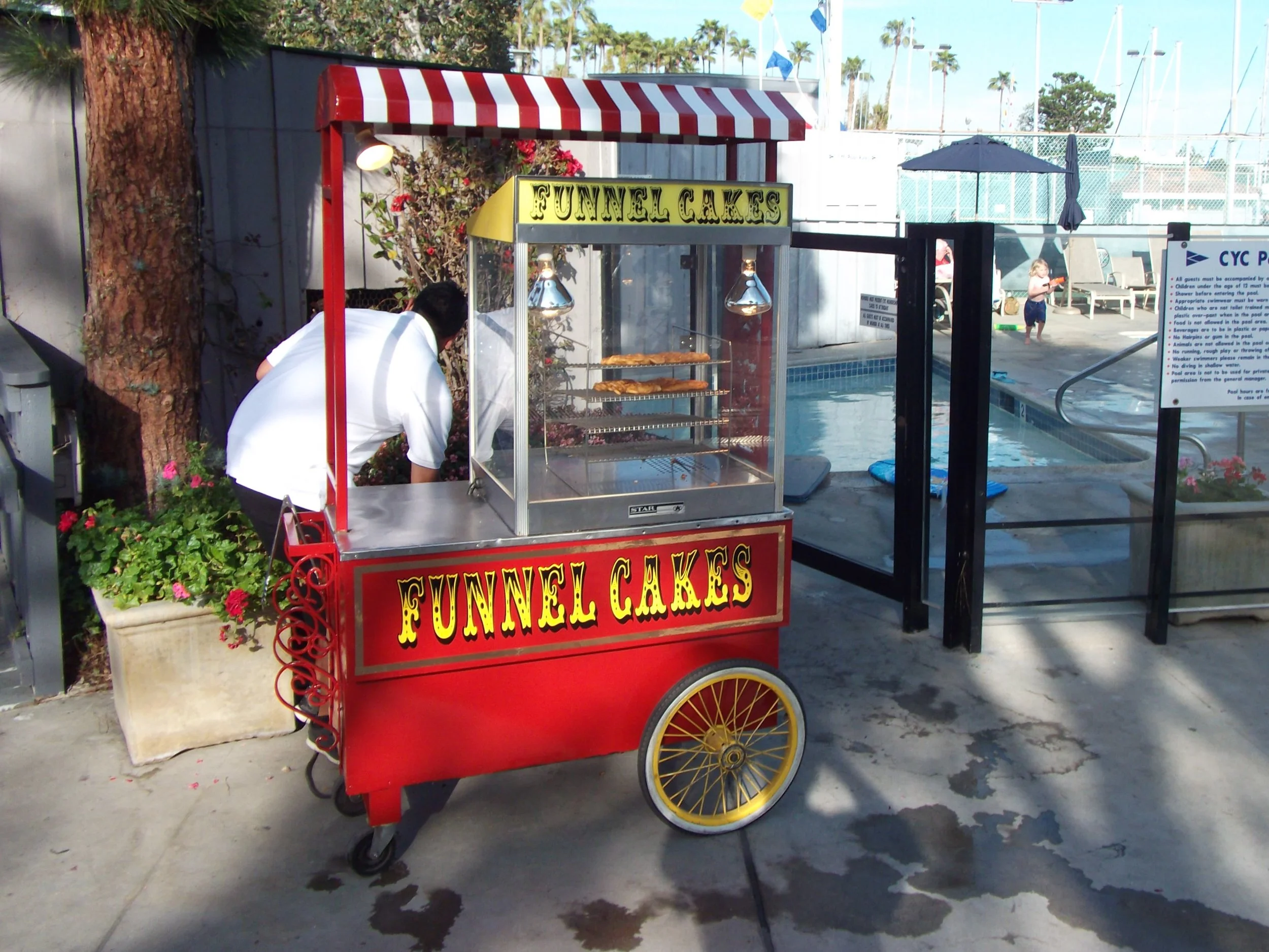 A red street cart with yellow wheels selling funnel cakes at an outdoor pool area, featuring a sign that says 'FUNNEL CAKES' and a striped red and white canopy.