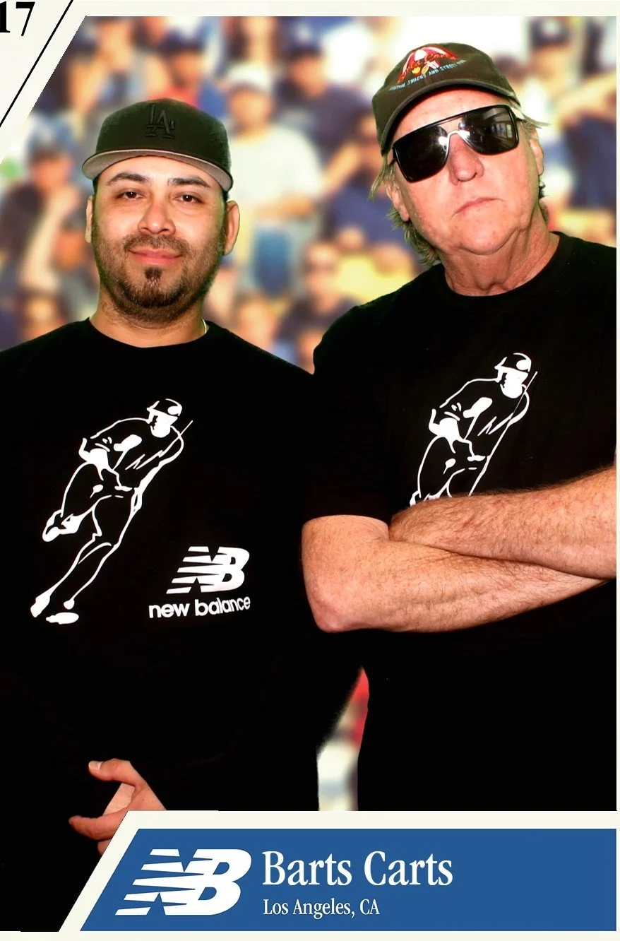 Two men wearing black T-shirts with a white running figure logo and 'New Balance' text. The man on the left is younger with a beard, baseball cap, and smiling. The man on the right is older, wearing sunglasses, a cap, and has arms crossed. Background