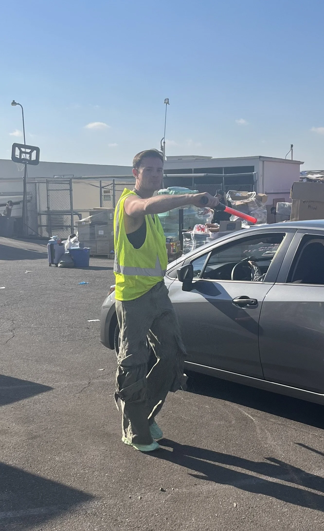A man in a yellow safety vest and beige cargo pants is playing with a pink toy lightsaber while standing beside a silver car in an outdoor parking lot or street. There are scattered items and a fenced area with boxes and bins behind him, under a clea