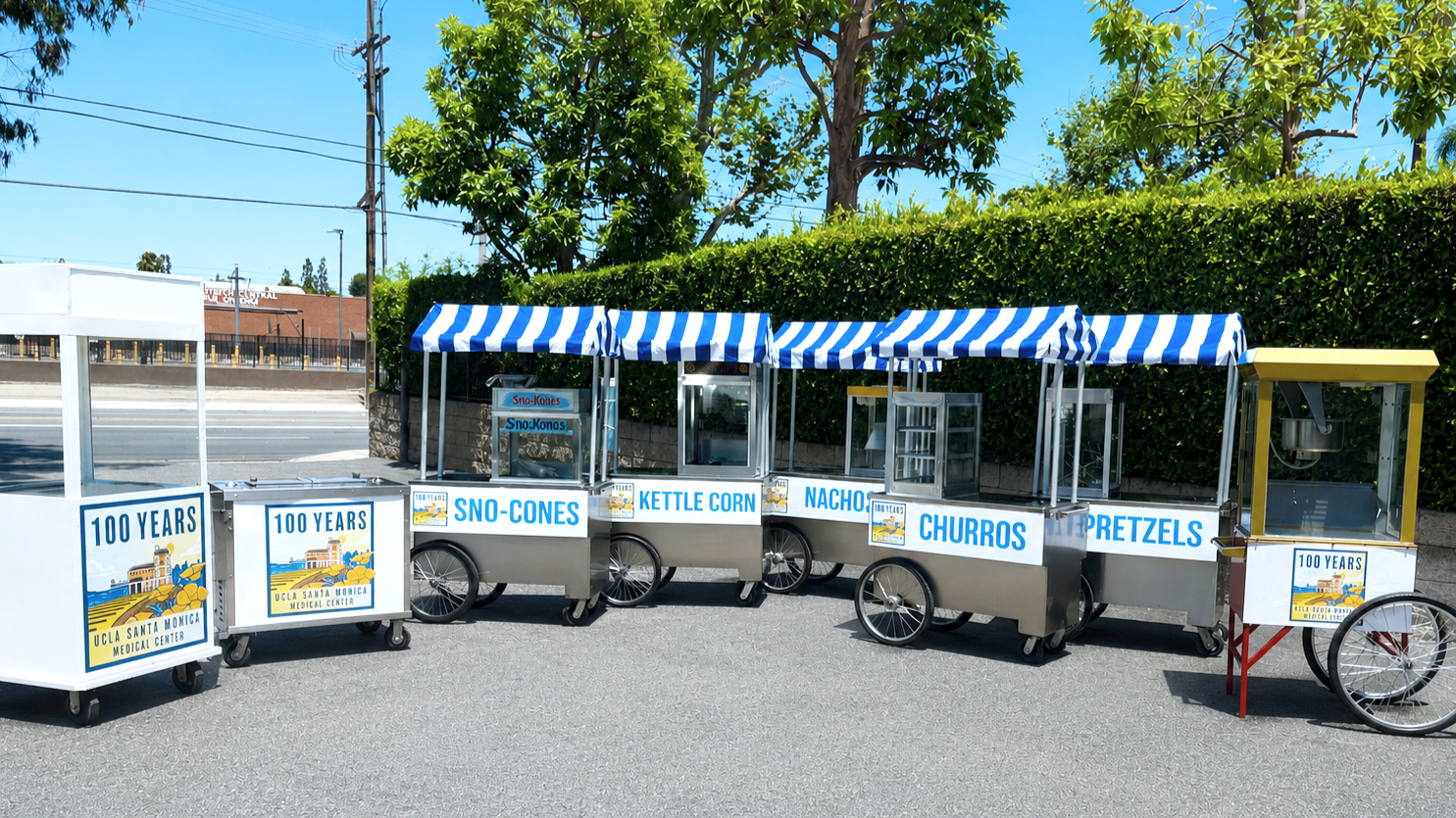 A row of vendor carts outside, each with a blue and white striped canopy, selling snacks like sno-cones, kettle corn, nachos, churros, pretzels, and cotton candy, with trees and a curb in the background.