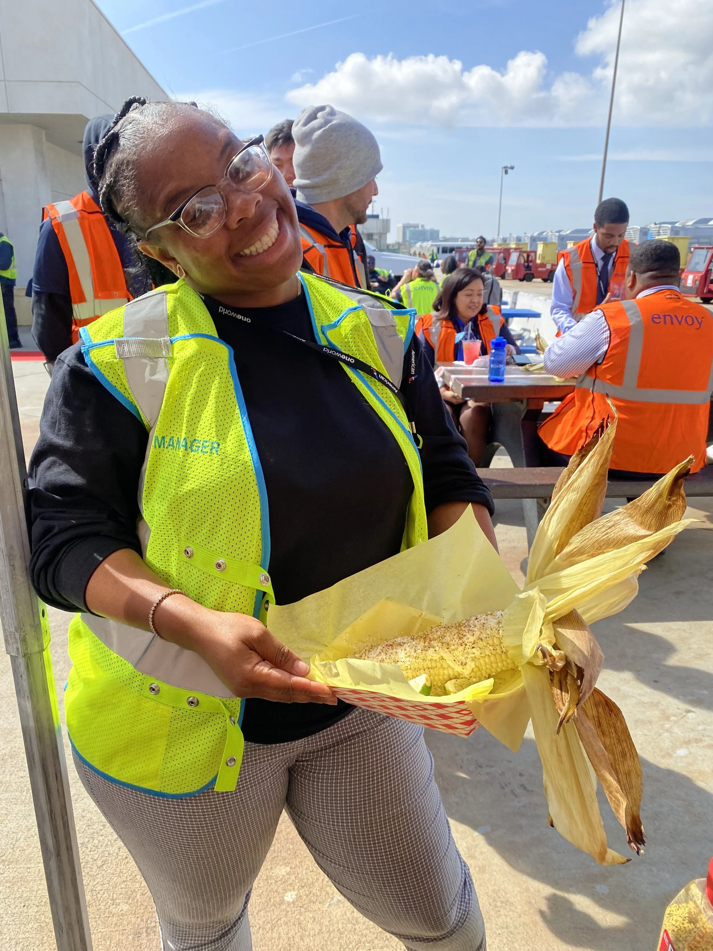 Woman smiling and holding a bouquet of corn on the cob wrapped in yellow paper while standing outdoors with people in safety vests and orange vests in the background.
