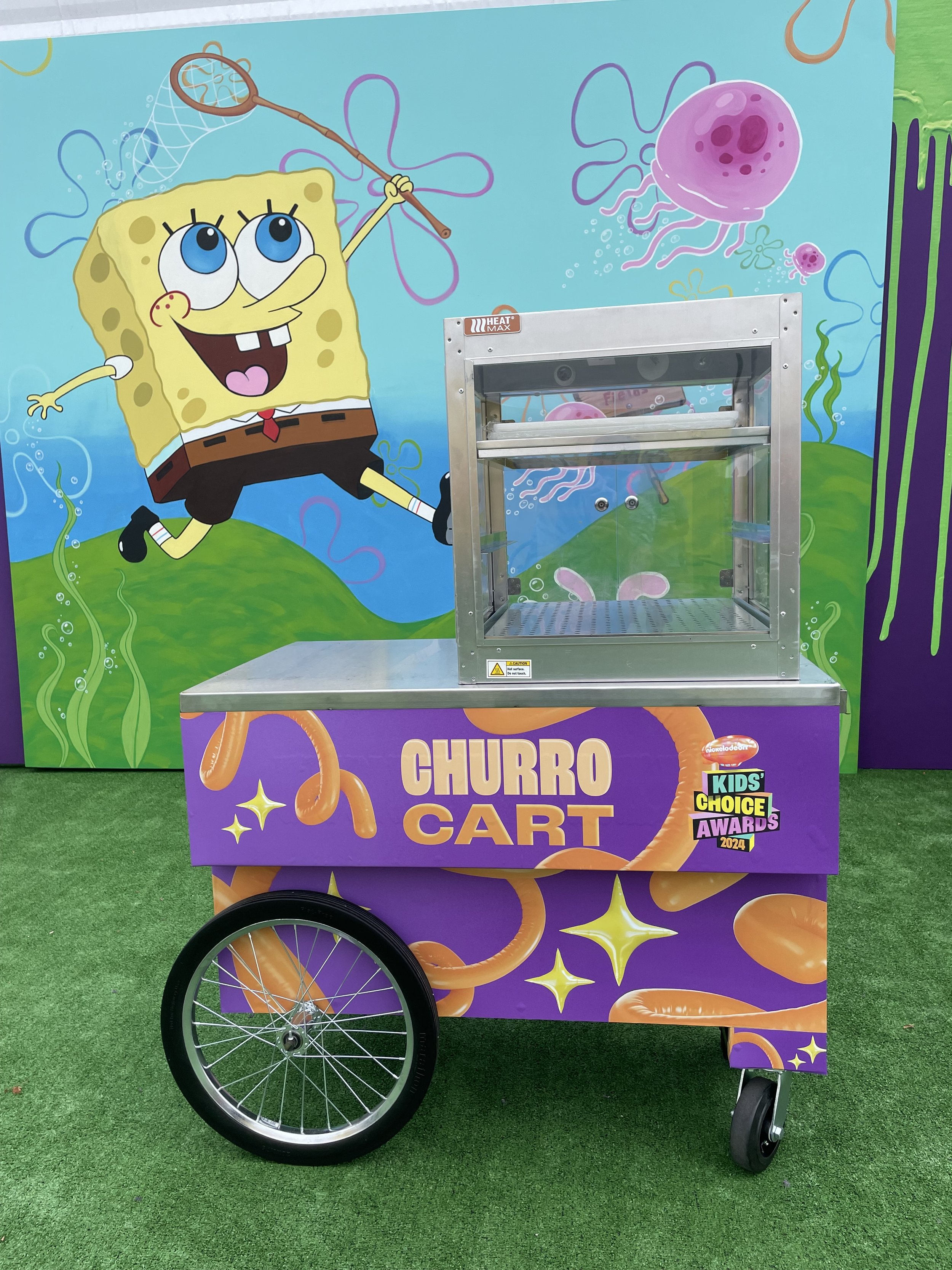 A colorful popcorn cart themed after SpongeBob SquarePants, featuring his cartoon character on a vibrant background with jellyfish and sea elements, with a metal frame and large wheel.