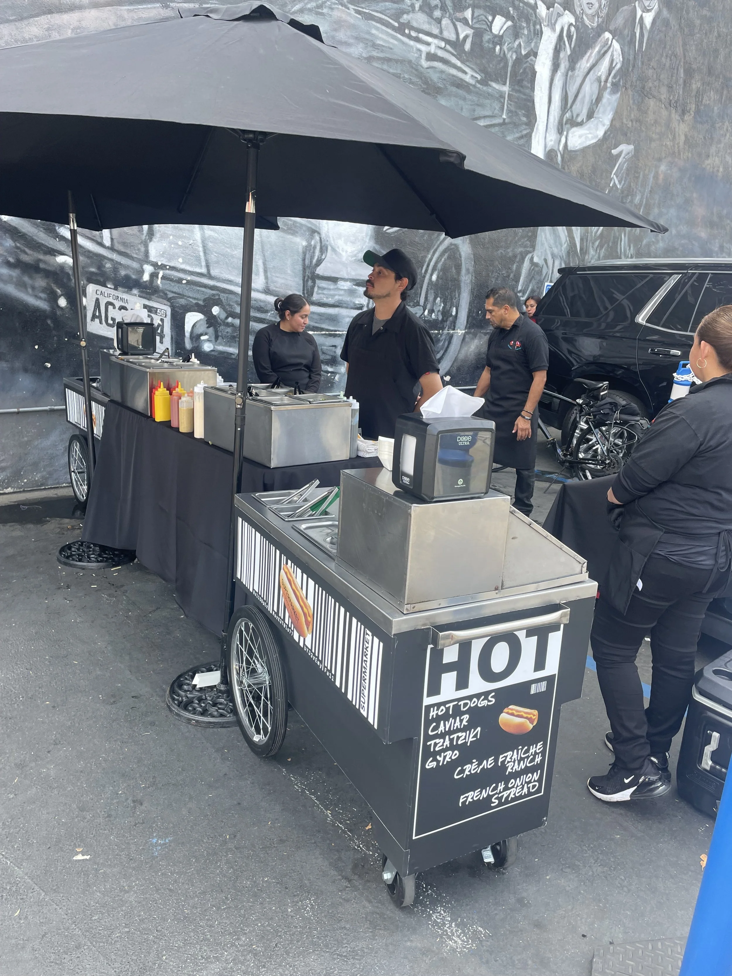 A food cart selling hot dogs and other snacks with a black canopy, located outdoors near a large mural. Several people are around the cart, and a woman is standing beside it. The cart has bottles of condiments and a chalkboard menu listing items such